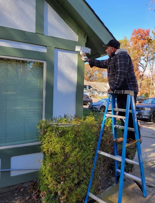 Man on ladder installs security light on a green and white house near a bush on a sunny day.