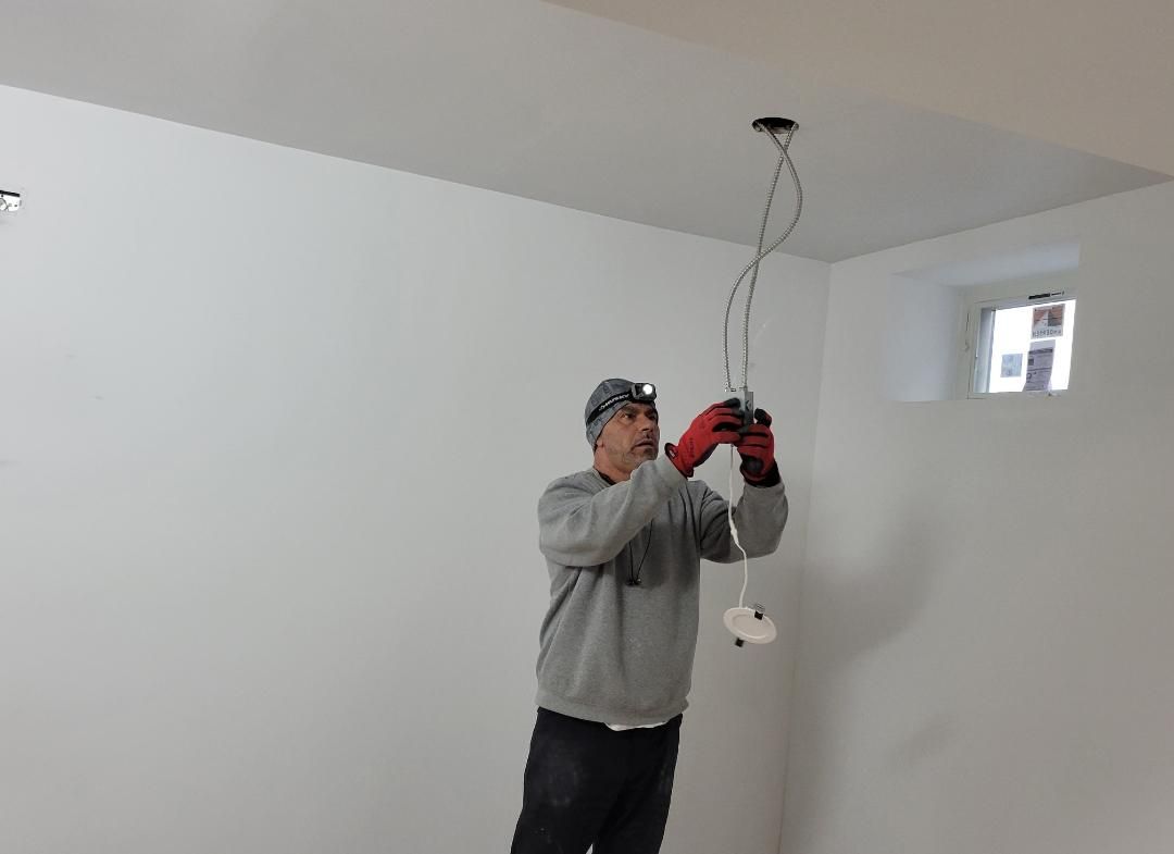 Man in work gloves, installing a light fixture in a white room, wearing a headlamp.