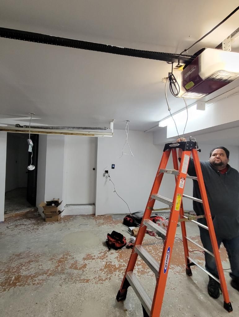 Man on ladder near garage door opener, looking up; tools on floor, white walls, and gray floor.