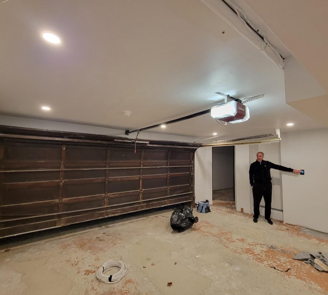Man stands in a garage, pointing towards a control panel. Garage door is closed, showing wooden panels.