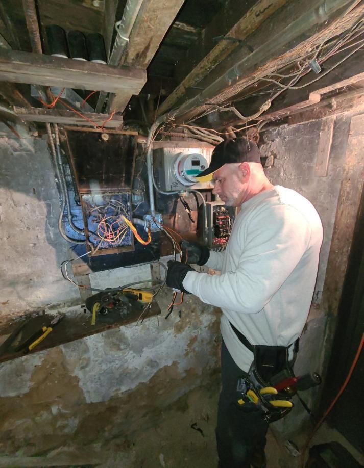 Electrician working on wires in a dimly lit basement, wearing gloves and a tool belt.