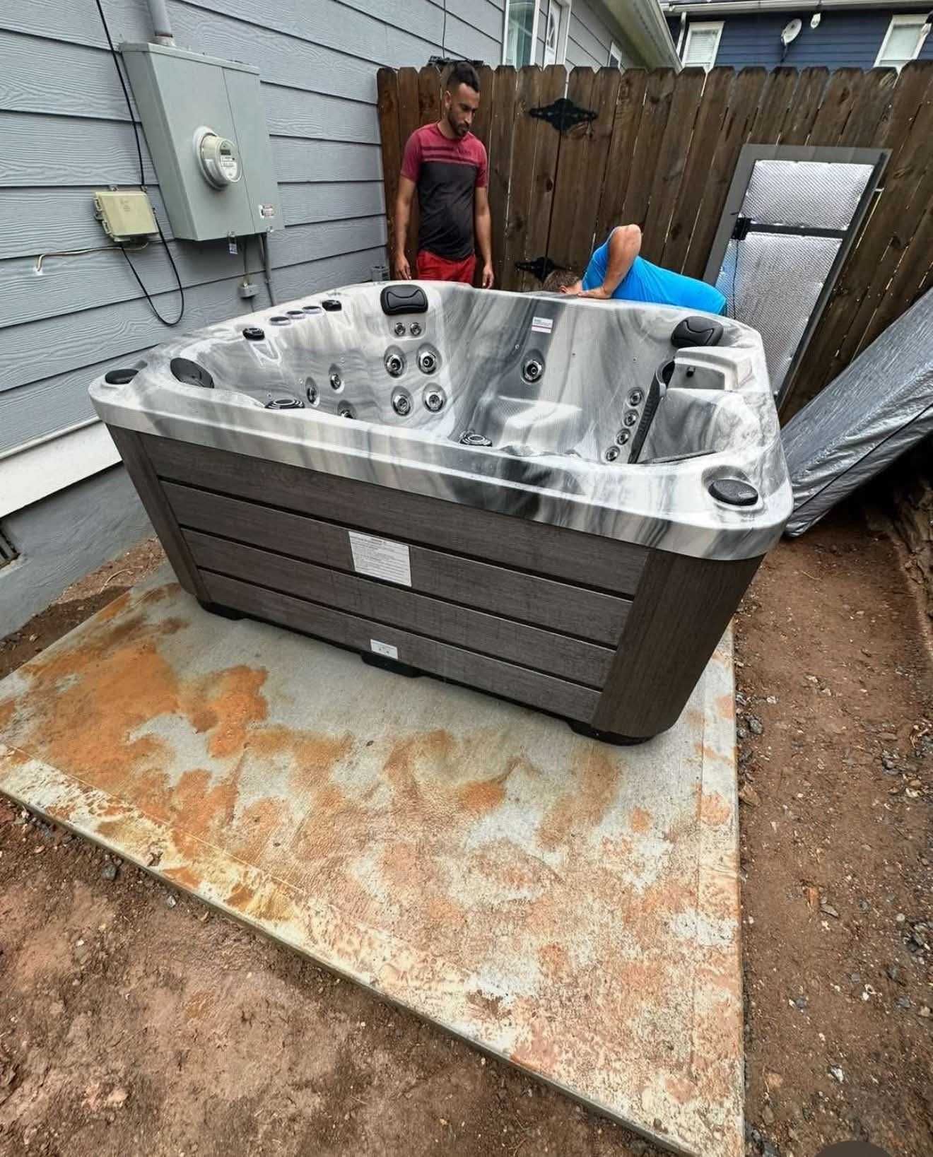 A new hot tub sits on a concrete pad in a backyard. Two men stand nearby; one is looking at the tub, the other is bending over.