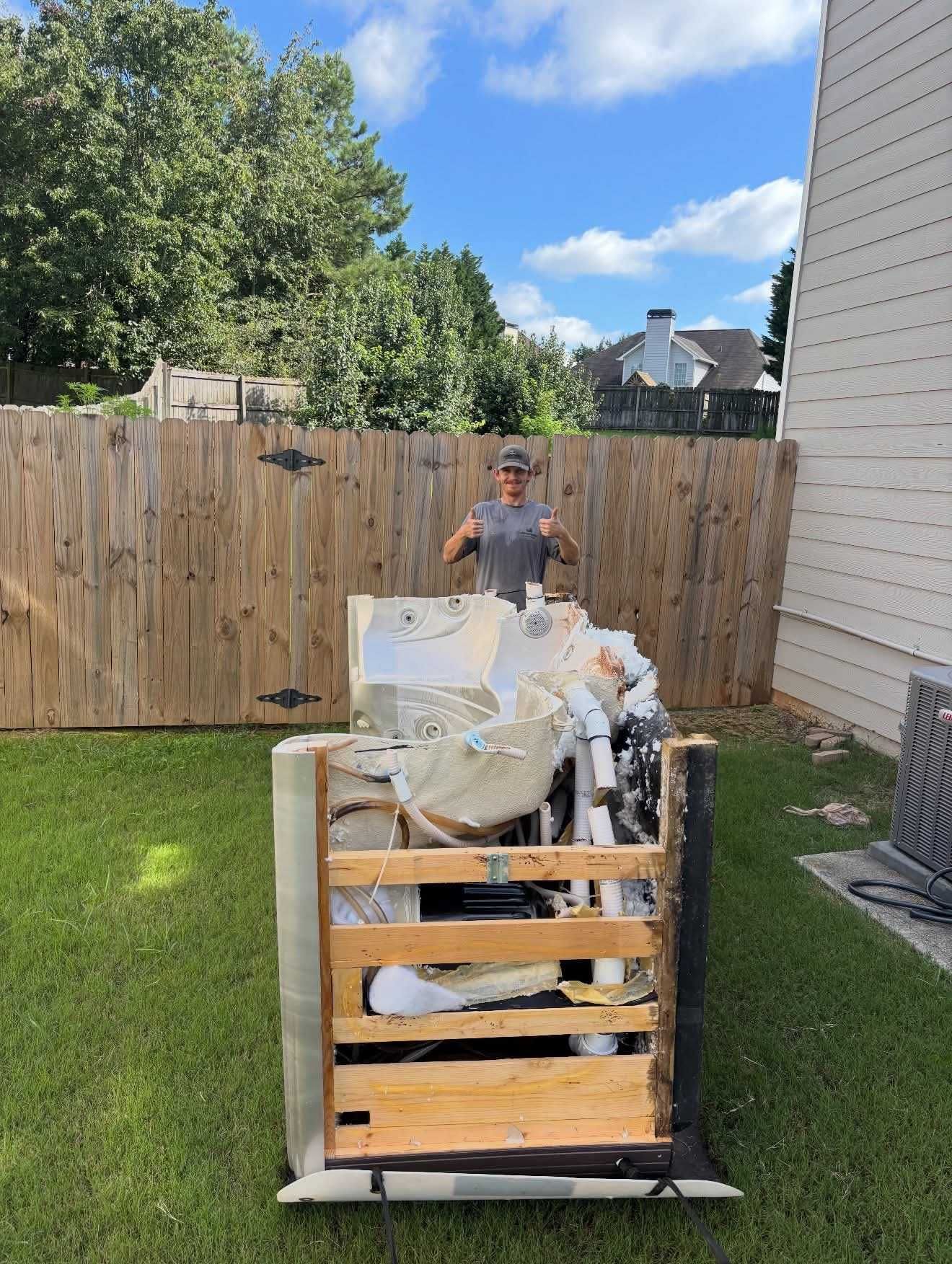 A person in a backyard gives a thumbs-up, standing behind a wooden bin overflowing with junk against a wooden fence.