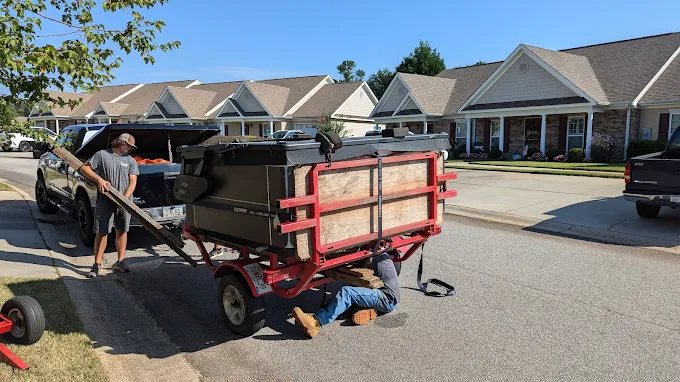 Two men working on a trailer in a residential street. One is under it, the other standing near a truck.