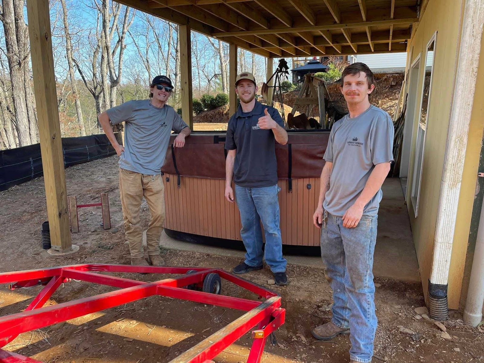 Three people standing by a small wooden structure under a roof, with a red trailer frame in the foreground.