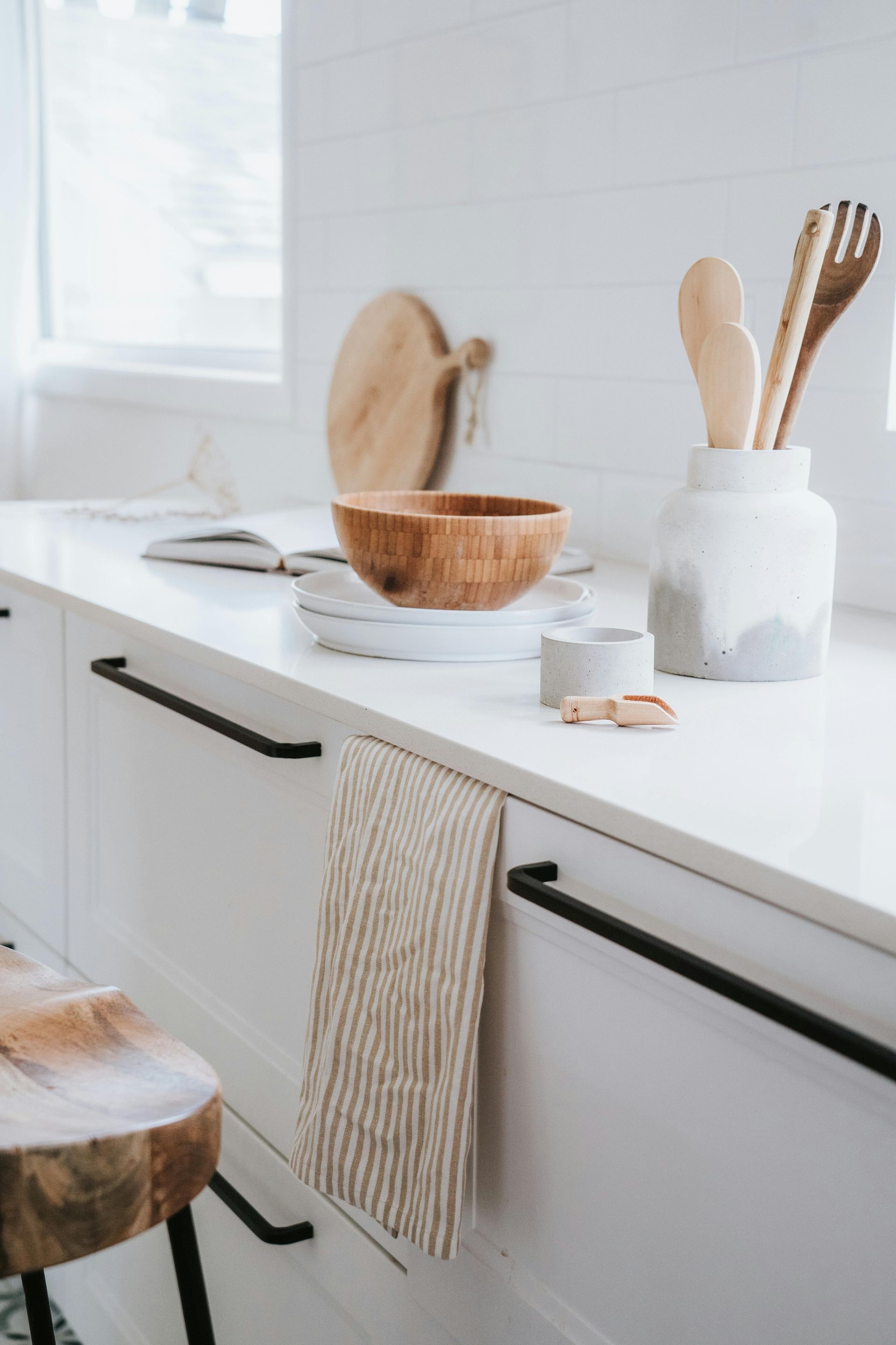 White kitchen counter with wooden utensils, bowl, and cutting board. A striped towel hangs below.