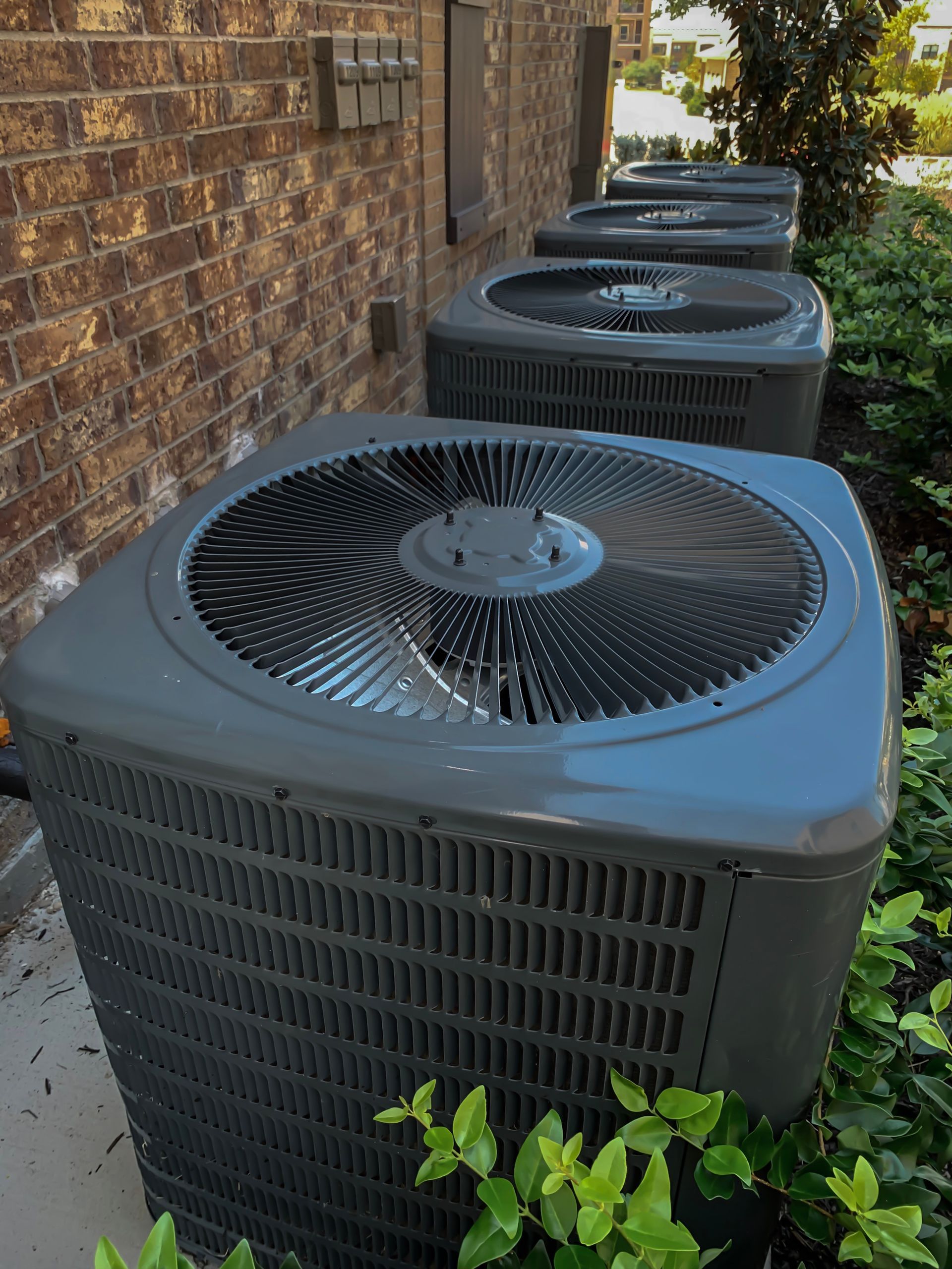 Row of gray air conditioning units next to a brick wall and bushes.