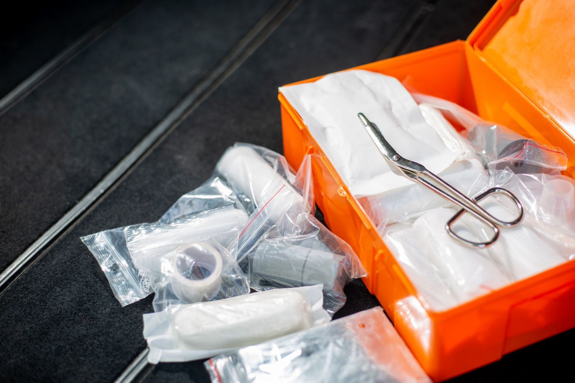 Orange first aid kit with contents scattered on black surface, including bandages and scissors.