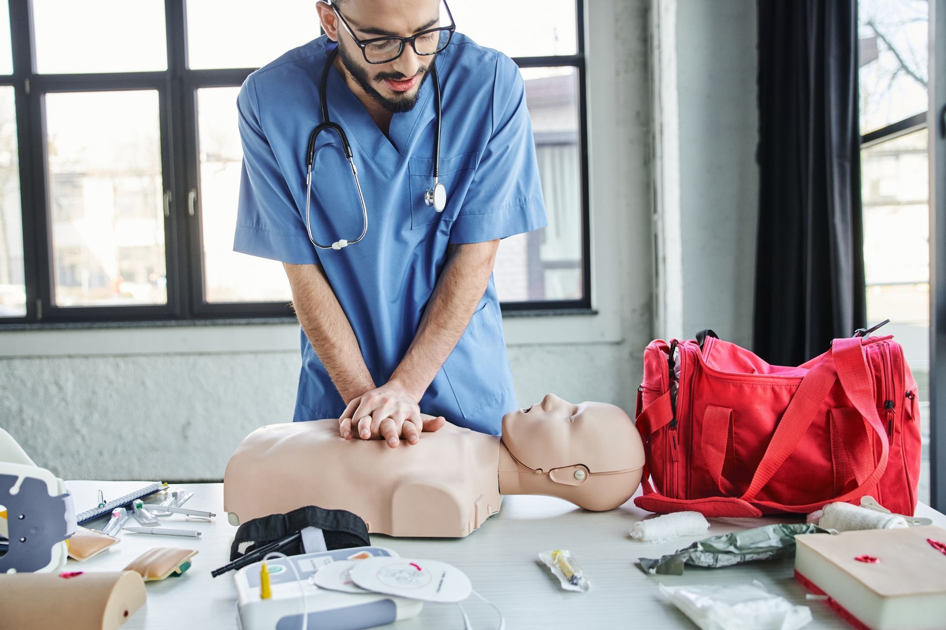 A person in scrubs practices CPR on a mannequin. Red medical bag sits on a table with medical supplies.