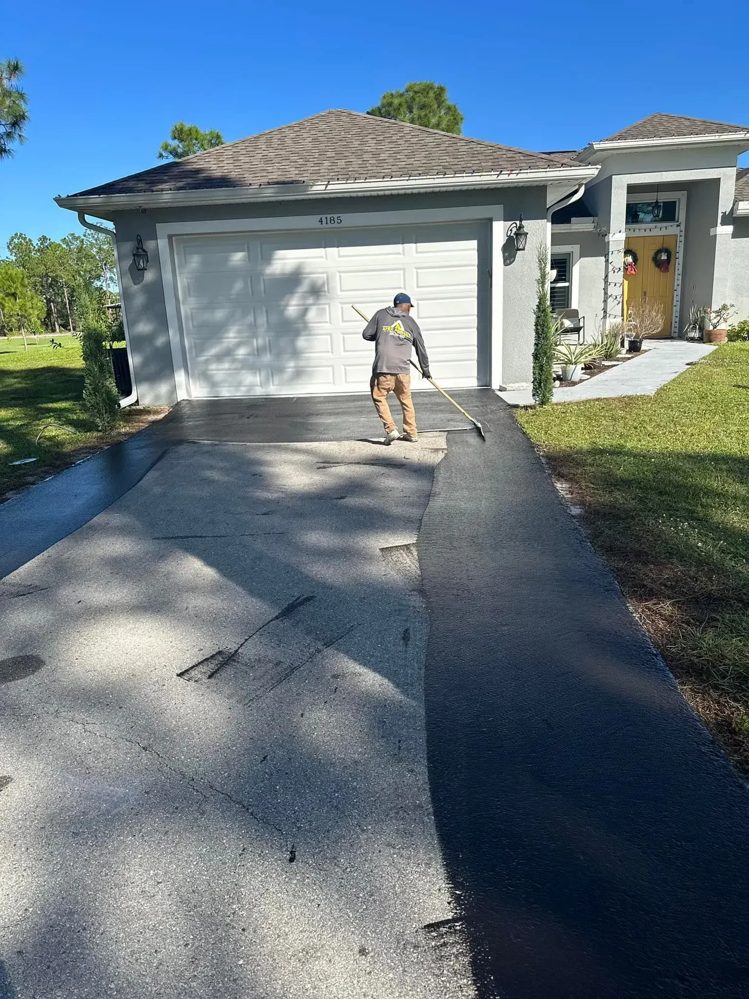 Person sealing a driveway in front of a gray house on a sunny day.