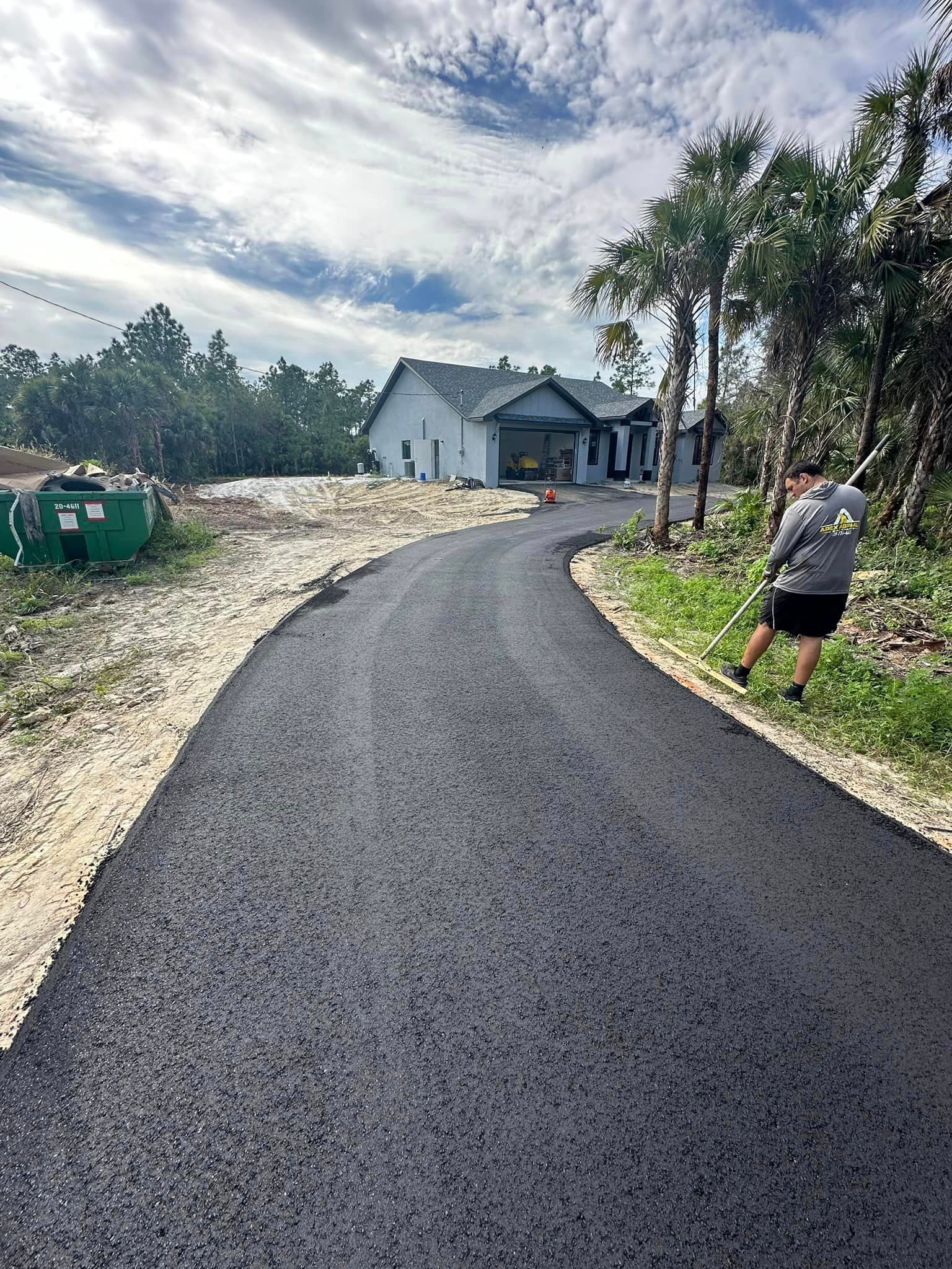 Asphalt driveway curves to a light-colored house. A person works on the right, clearing brush.