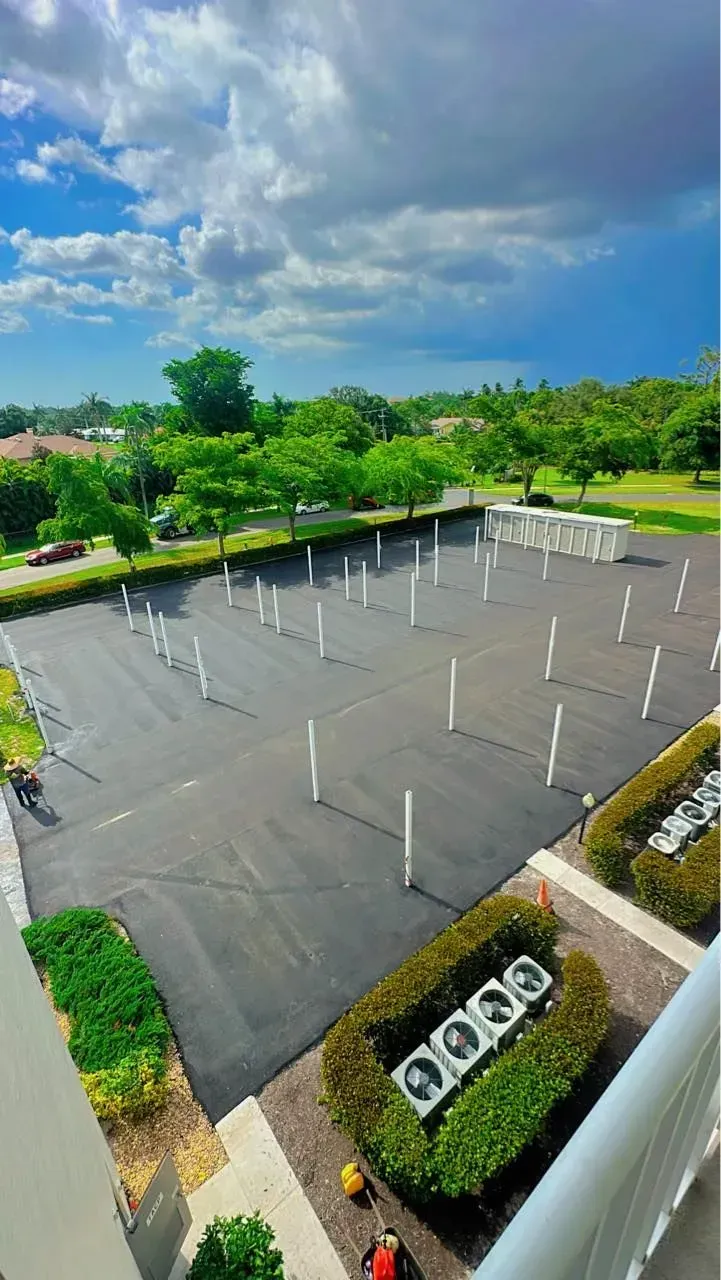 Asphalt parking lot with white poles. Green bushes and trees surround the lot under a cloudy blue sky.