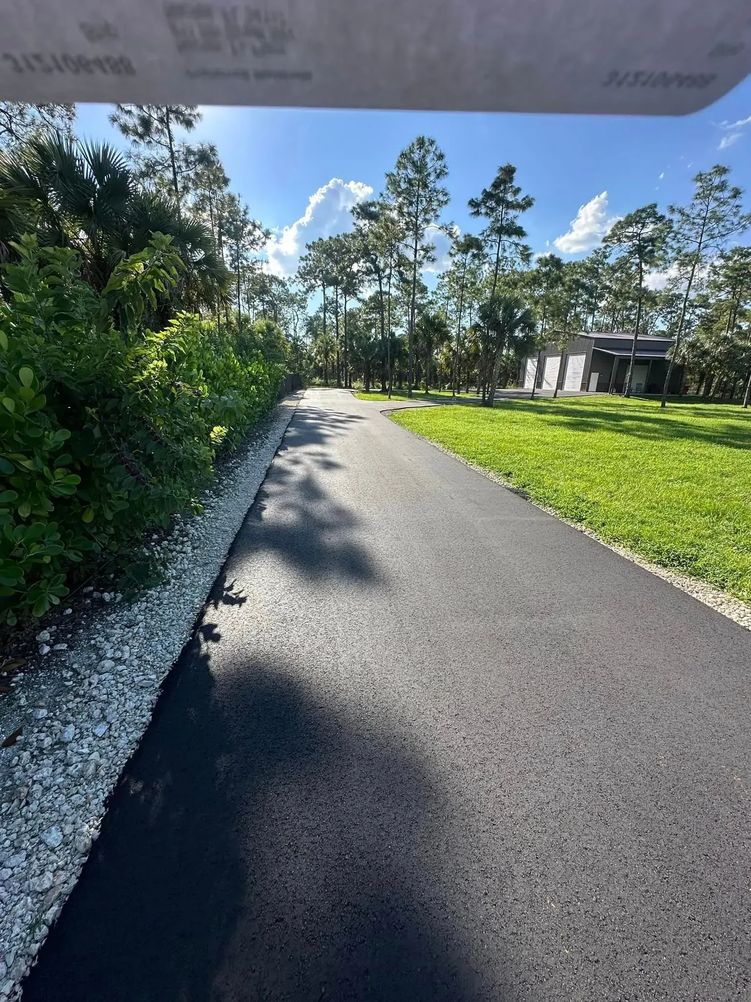 Paved path through trees, green grass, and white gravel. Bright sky with fluffy clouds.