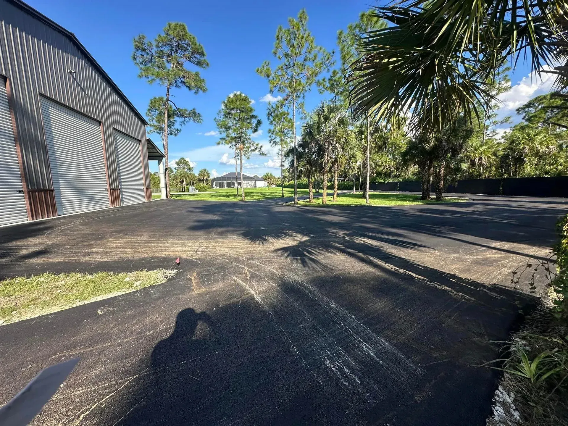 Asphalt lot in front of a metal building and trees, with a blue sky and shadows.