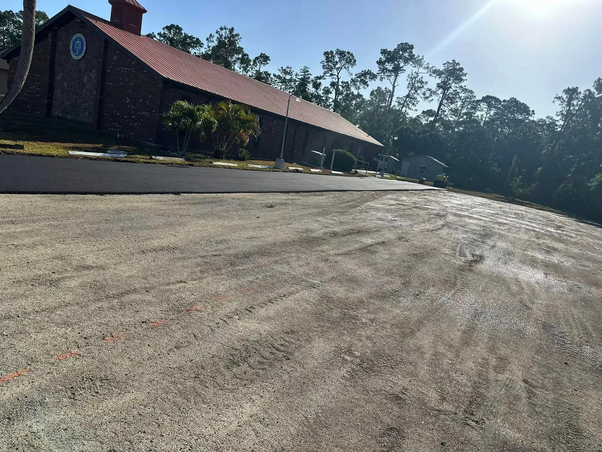 Gravel parking area slopes down toward a red brick building with a sloped tile roof. Trees in background.