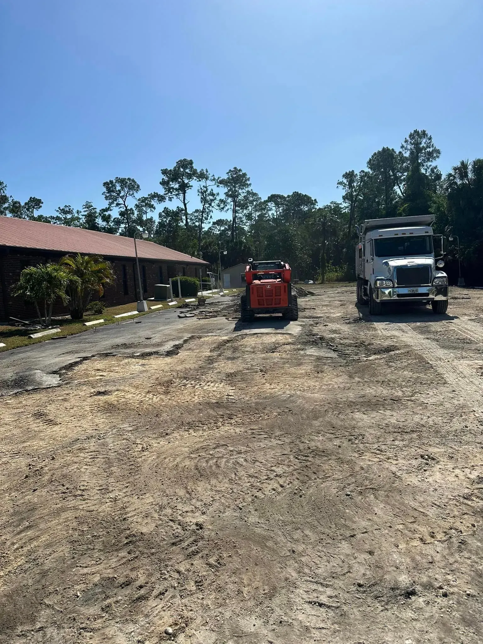 Construction site: Skid steer and dump truck on dirt road. Building and trees in the background under blue sky.