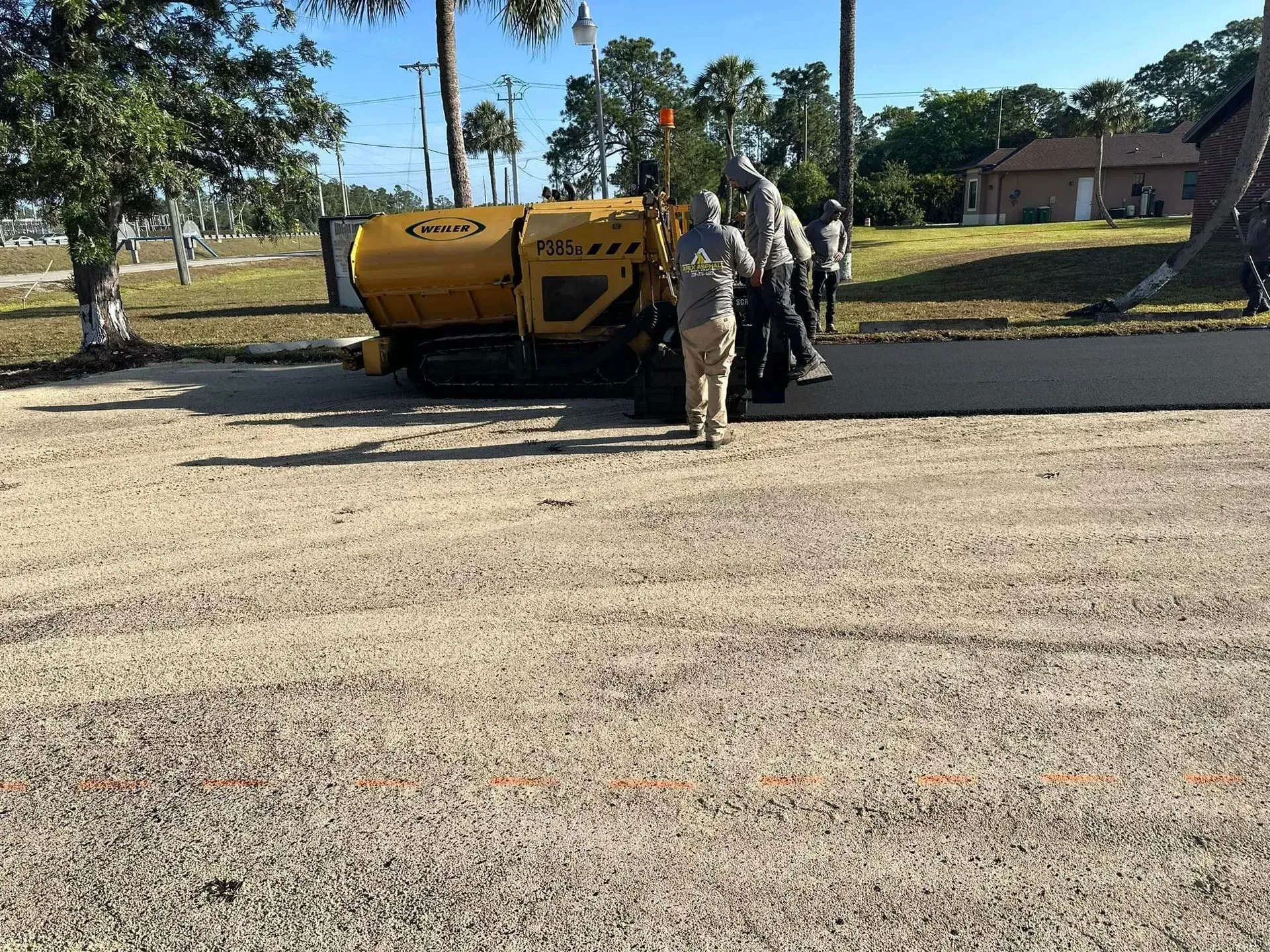 Asphalt paving machine laying asphalt in a gravel lot with crew members standing nearby.