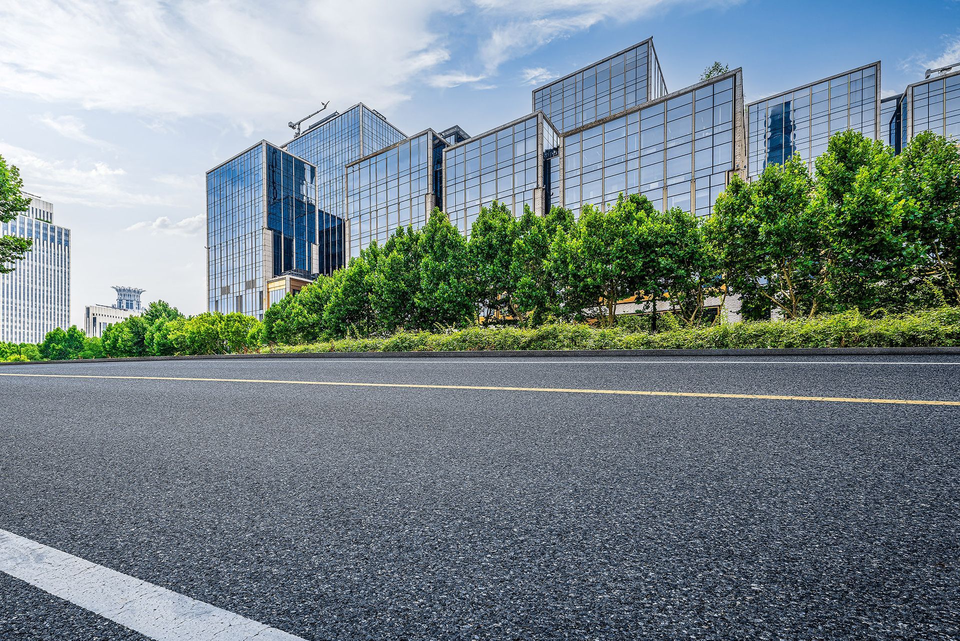 Asphalt road with yellow lines, lined with green trees, leading towards modern glass buildings under a blue sky.