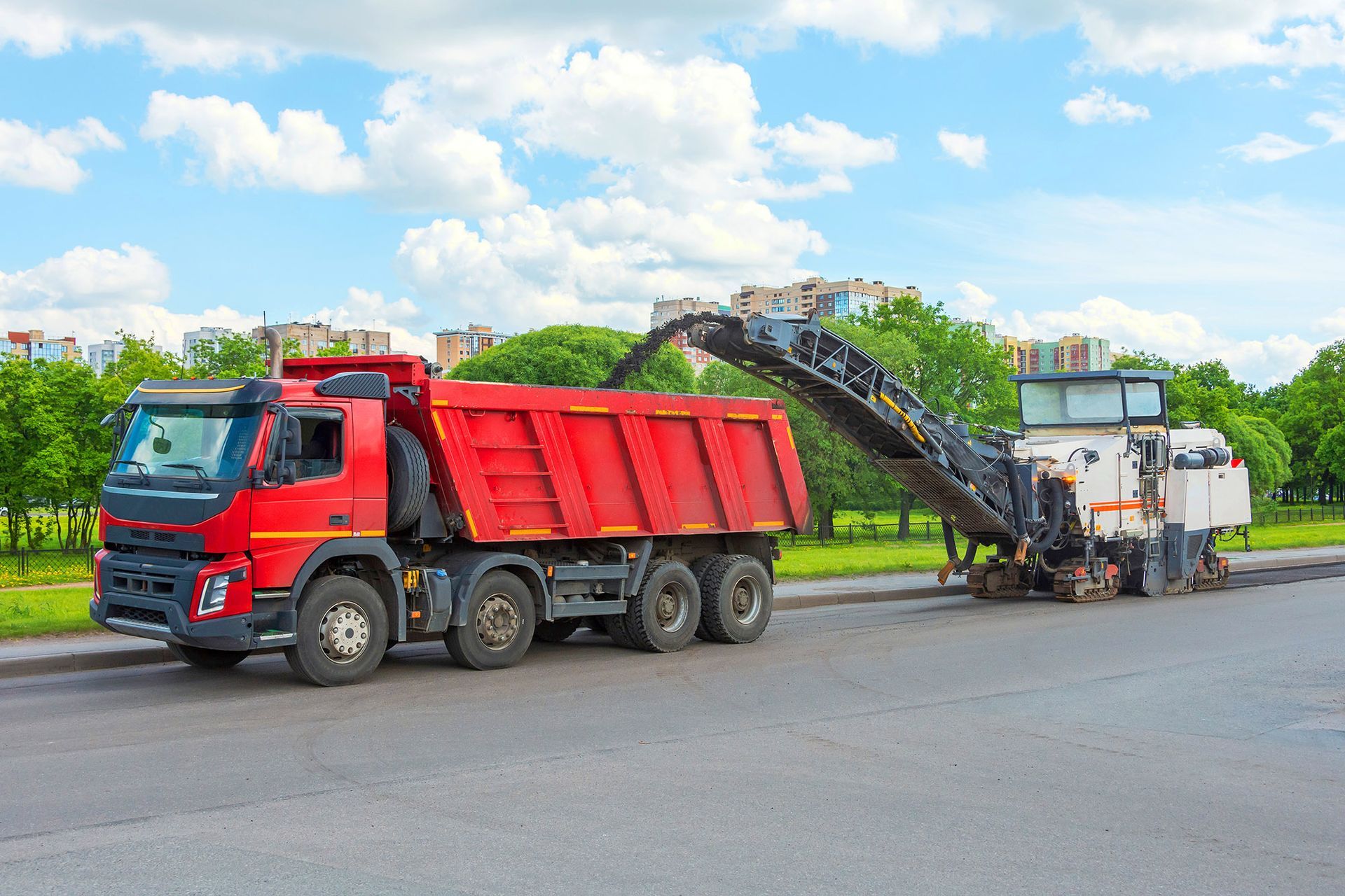 Red dump truck being loaded with asphalt by a milling machine on a city street.
