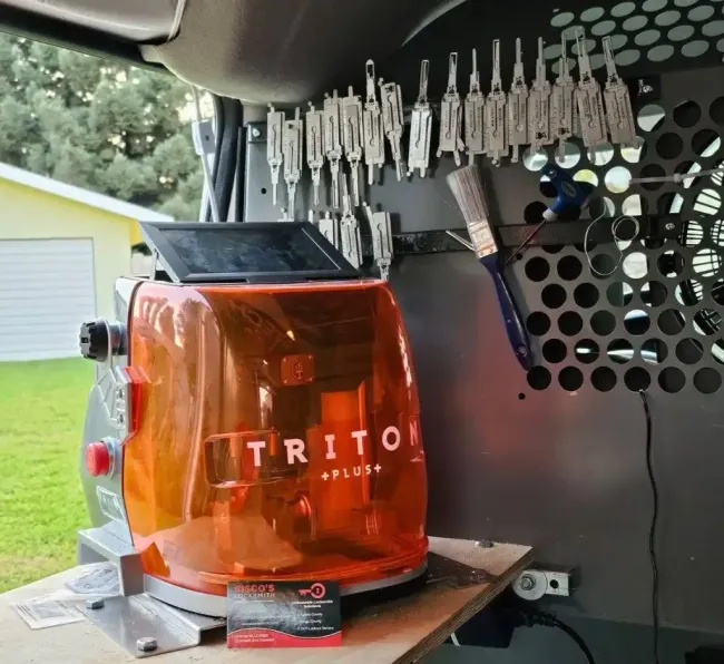 An orange Triton Plus key cutting machine sits on a workbench with a rack of keys hanging on a perforated metal wall.