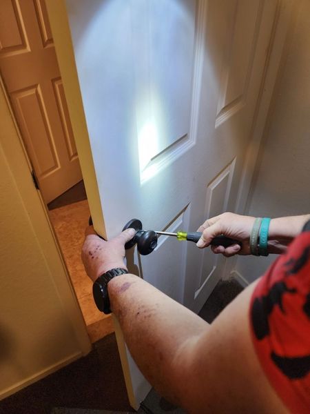 A person uses a screwdriver to remove a round, dark-colored door knob from a white interior door.