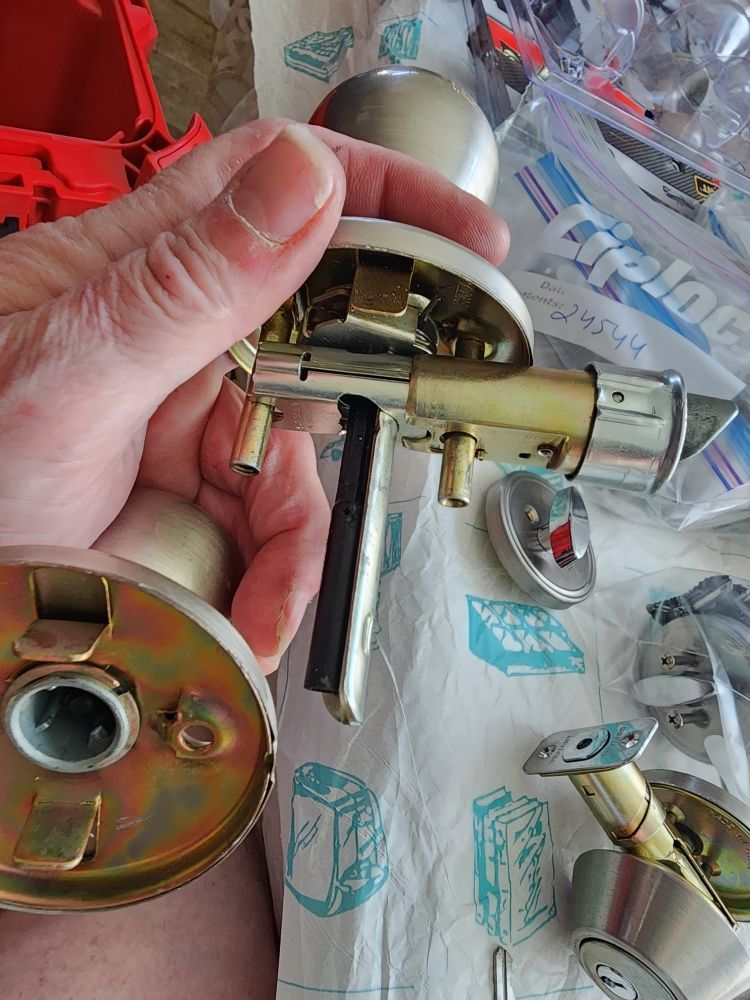 A hand holds a disassembled brass door lock mechanism with internal components visible, resting on paper near other parts.