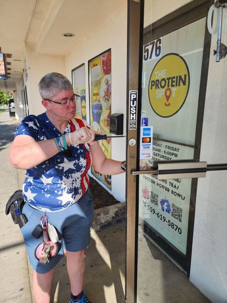 A person wearing a patriotic shirt uses a screwdriver to work on the door lock of a shop called The Protein Spot.