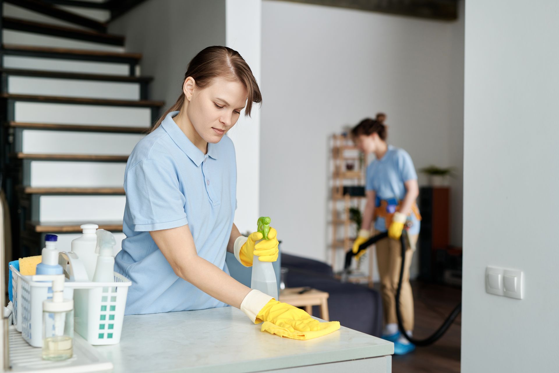 Two people cleaning a home: one spraying a surface, the other vacuuming. Brightly lit interior.