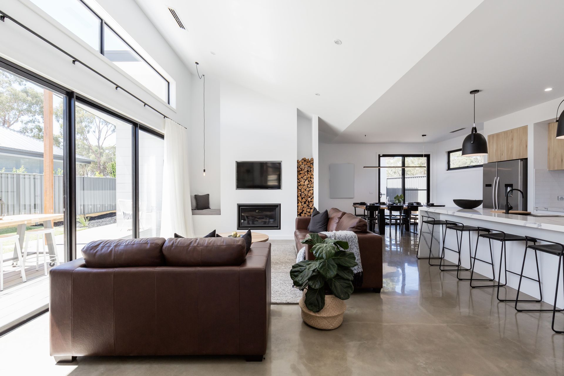 Modern open-plan living room with brown leather sofa, fireplace, and large windows overlooking backyard.