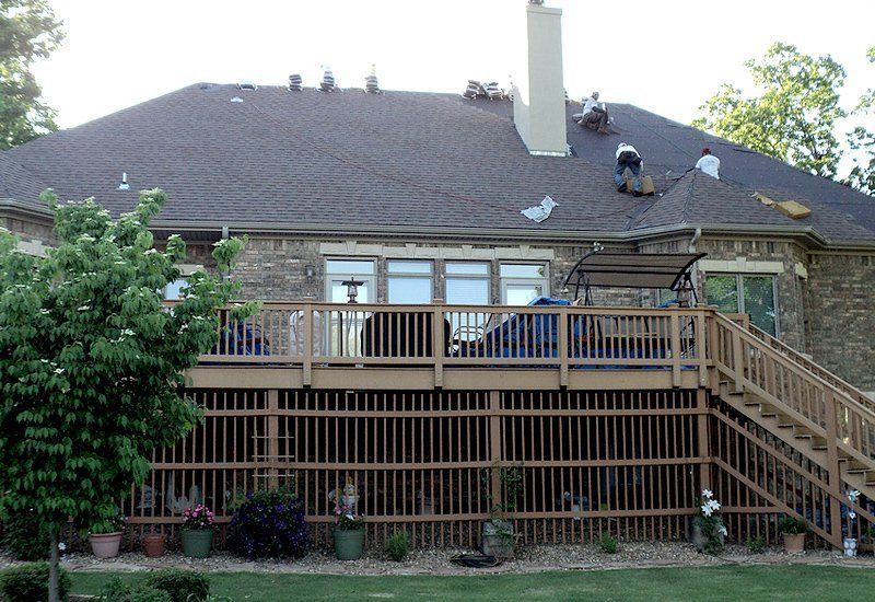 Roofers working on a dark shingled roof of a brick house with a wooden deck and a lush lawn.