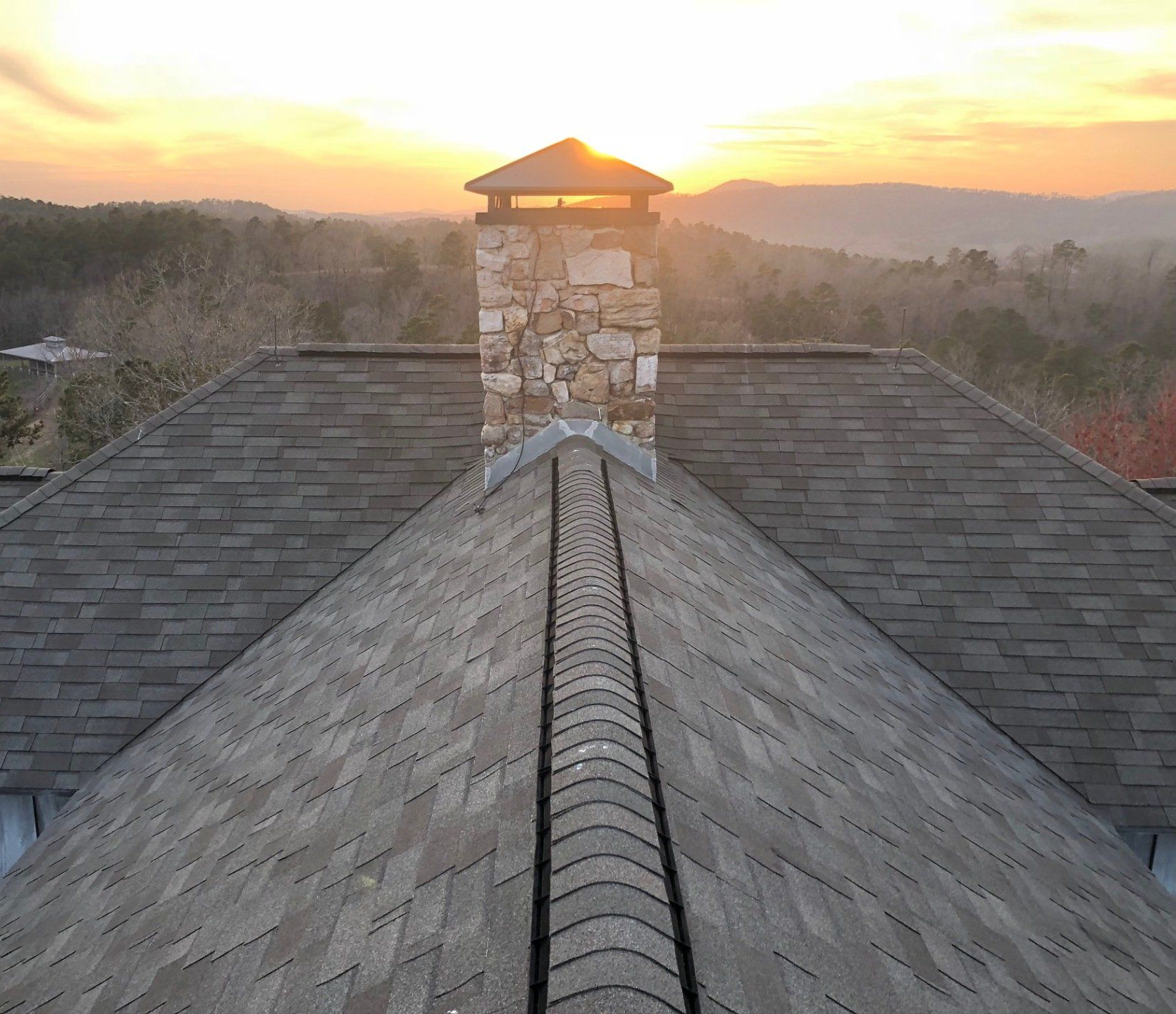 Rooftop with chimney at sunset; view of mountains in the distance, roof shingles are gray.
