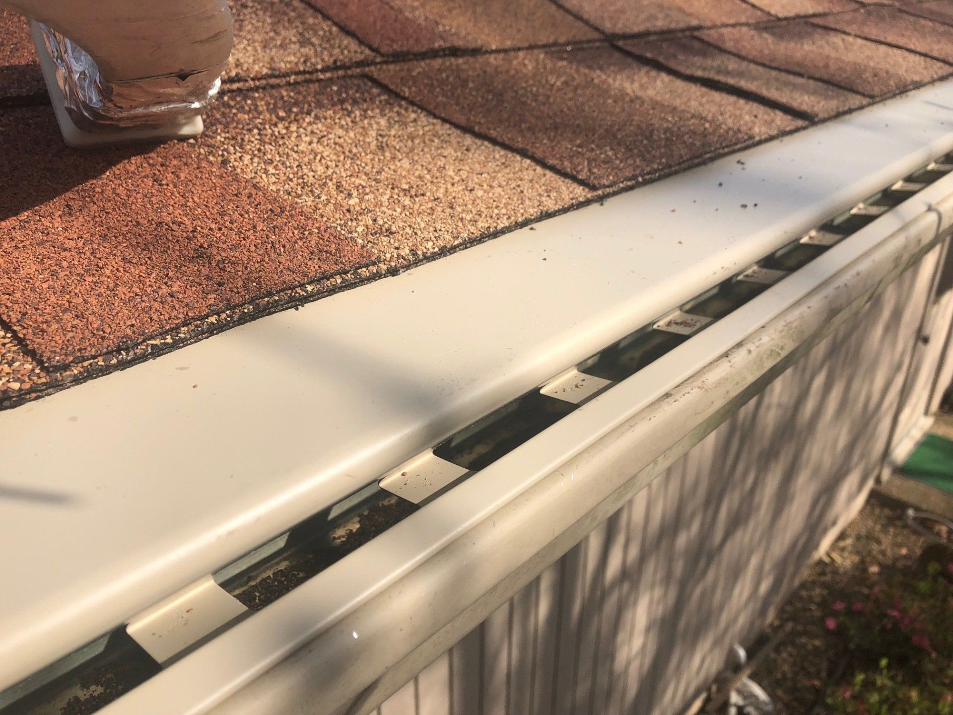 White gutter on a house roof. Debris visible inside gutter near brown roof shingles.