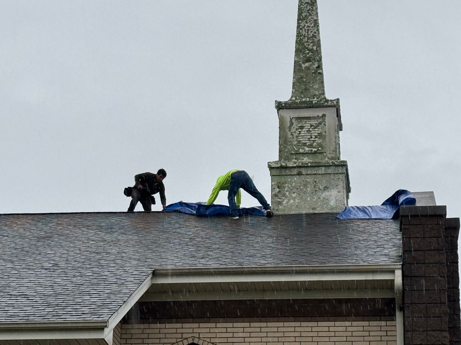 Two workers on a church roof, covering it with a blue tarp. Rainy weather.