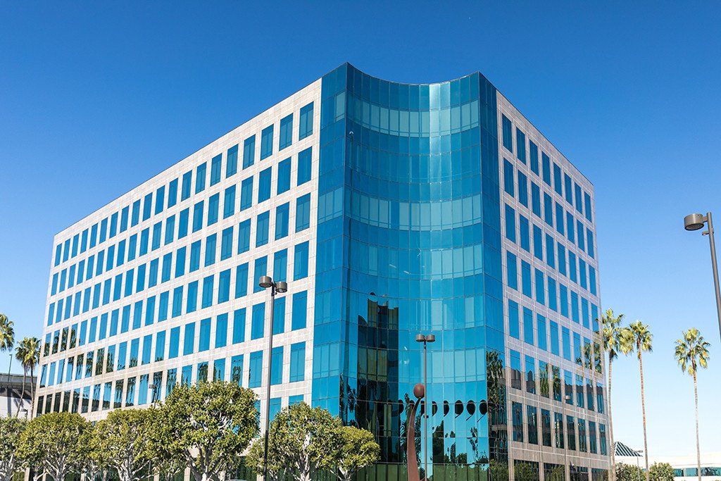 Modern glass office building under a blue sky, with palm trees lining the street.