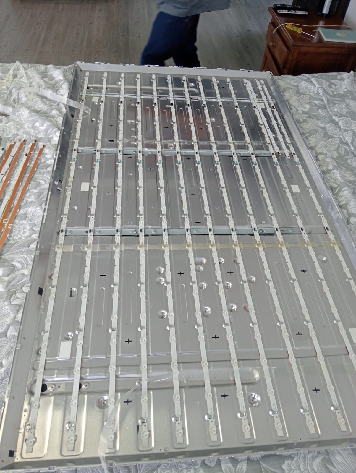 Panel de TV LED retroiluminado con tiras de luz, probablemente para reparación. Marco plateado con persona visible.