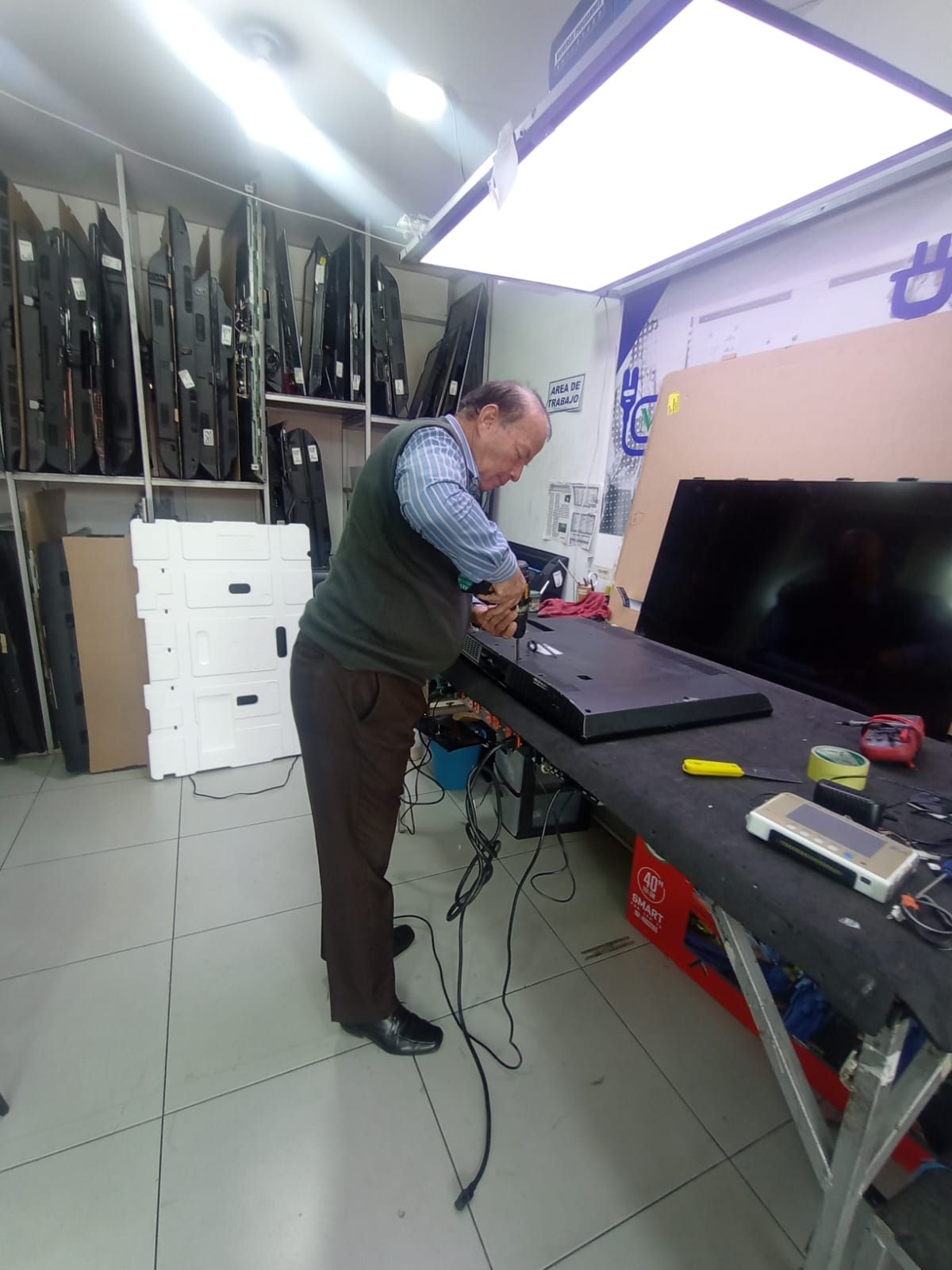 Hombre reparando un televisor en un taller, rodeado de piezas y herramientas, iluminado por una luz cenital.