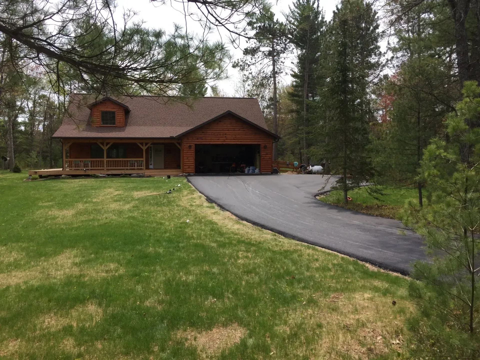 A brown wooden house with a shingled roof, a front porch, and an attached garage sits at the end of a paved driveway.