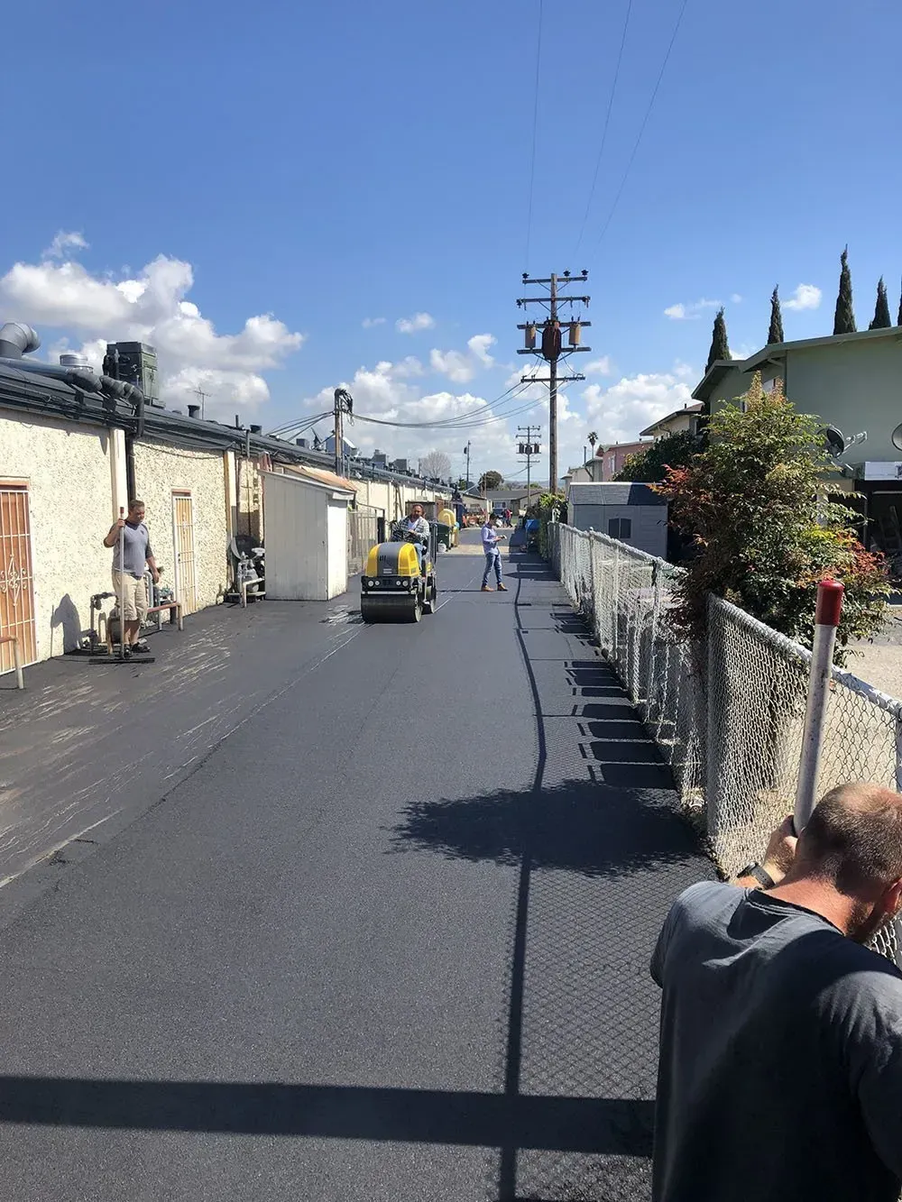 Workers operate a roller on a newly paved alley between a wall and a chain-link fence under a clear blue sky.