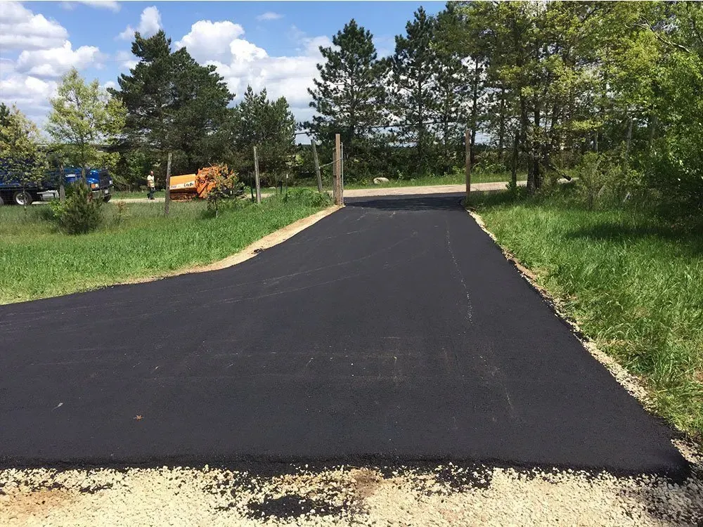 A freshly paved, black asphalt driveway extending from a gravel area toward a row of trees under a blue, cloudy sky.
