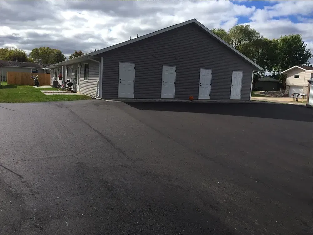 A grey residential building with four white exterior doors facing a large, freshly paved asphalt parking lot.