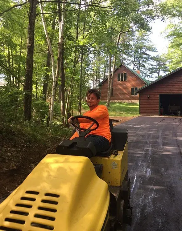A person wearing an orange shirt sits on a yellow construction roller on a paved driveway near a log cabin in the woods.