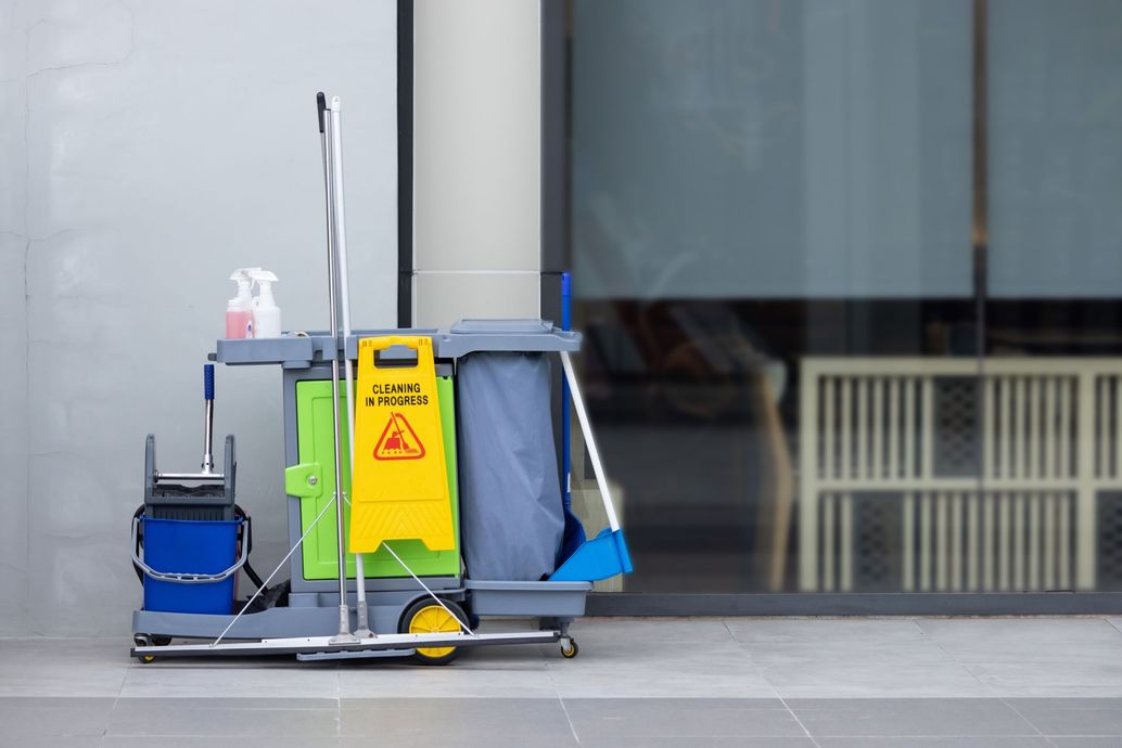 A custodial cleaning cart with a yellow warning sign, cleaning supplies, and a broom, standing on a tiled floor.