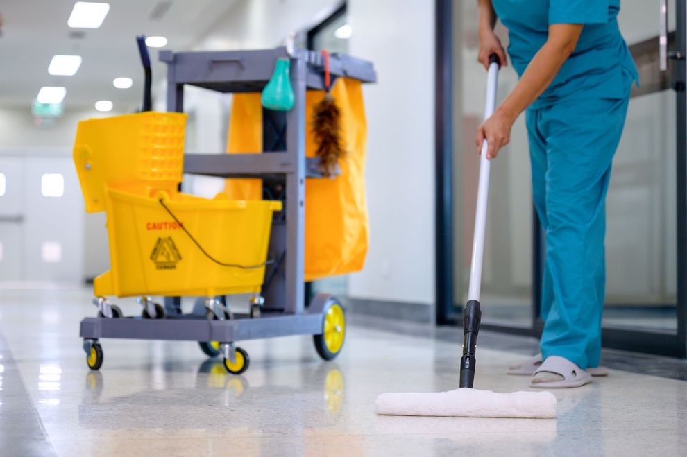 A person in teal scrubs mops a white tiled hospital hallway floor next to a yellow cleaning cart.