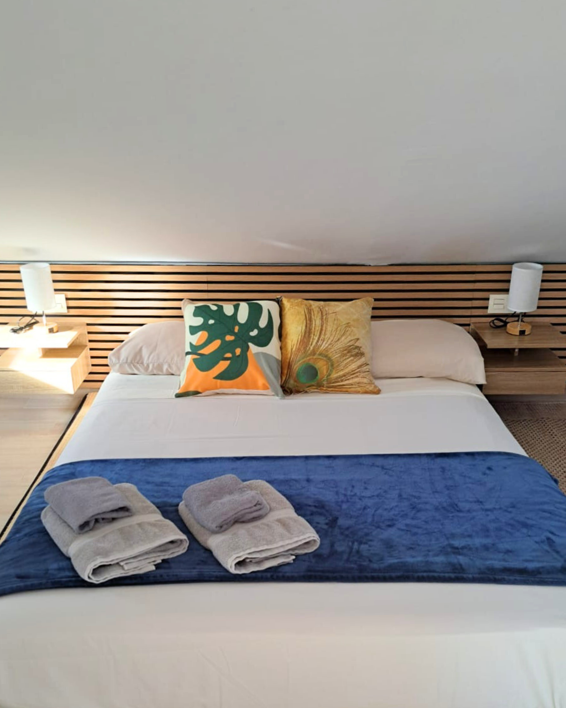 A neatly made bed with a white comforter, a navy blue blanket, grey towels, and two decorative pillows against a slat wall.
