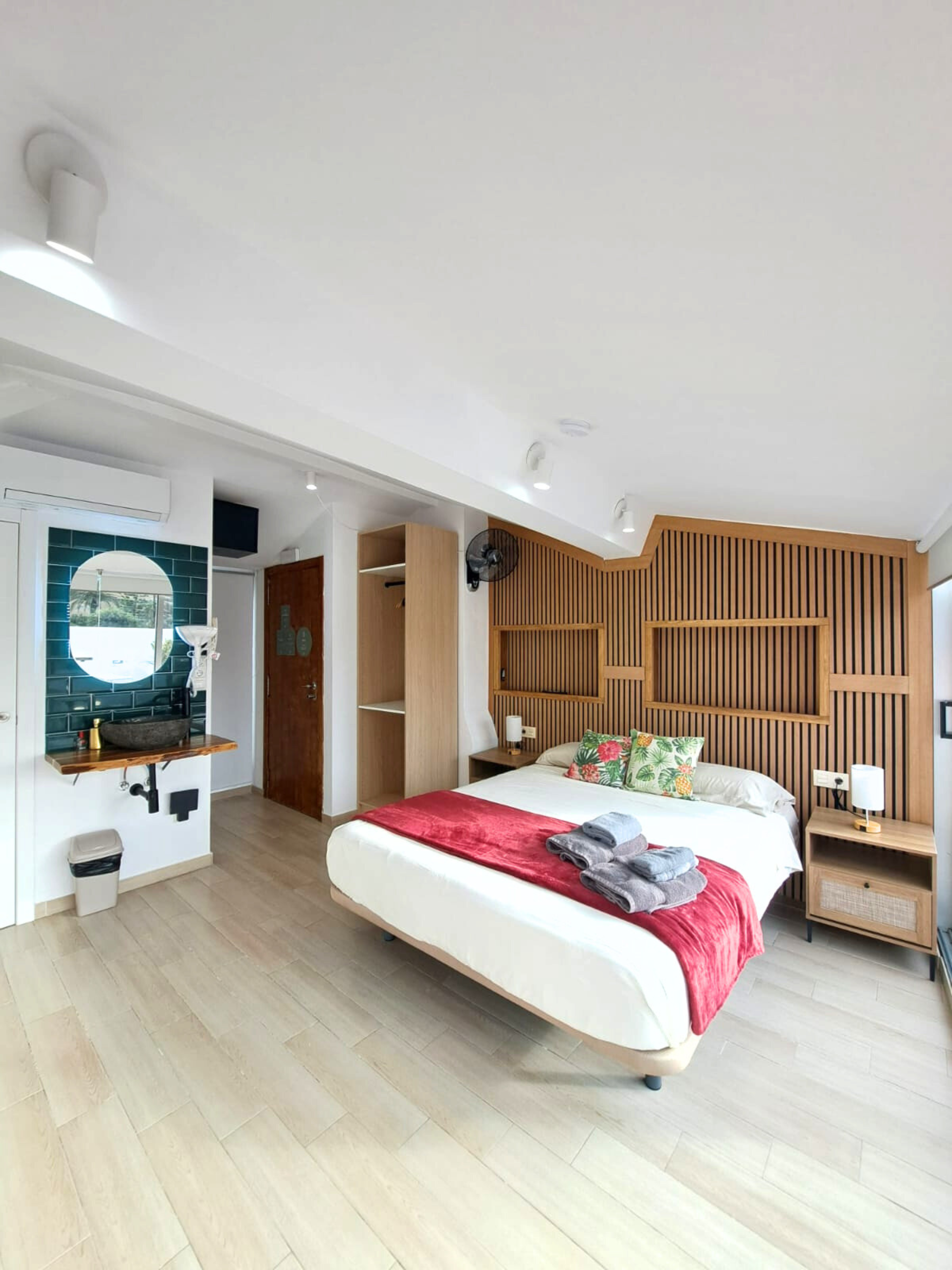 A modern hotel room with a bed featuring a red runner, wooden wall panels, a corner vanity, and a wooden closet.
