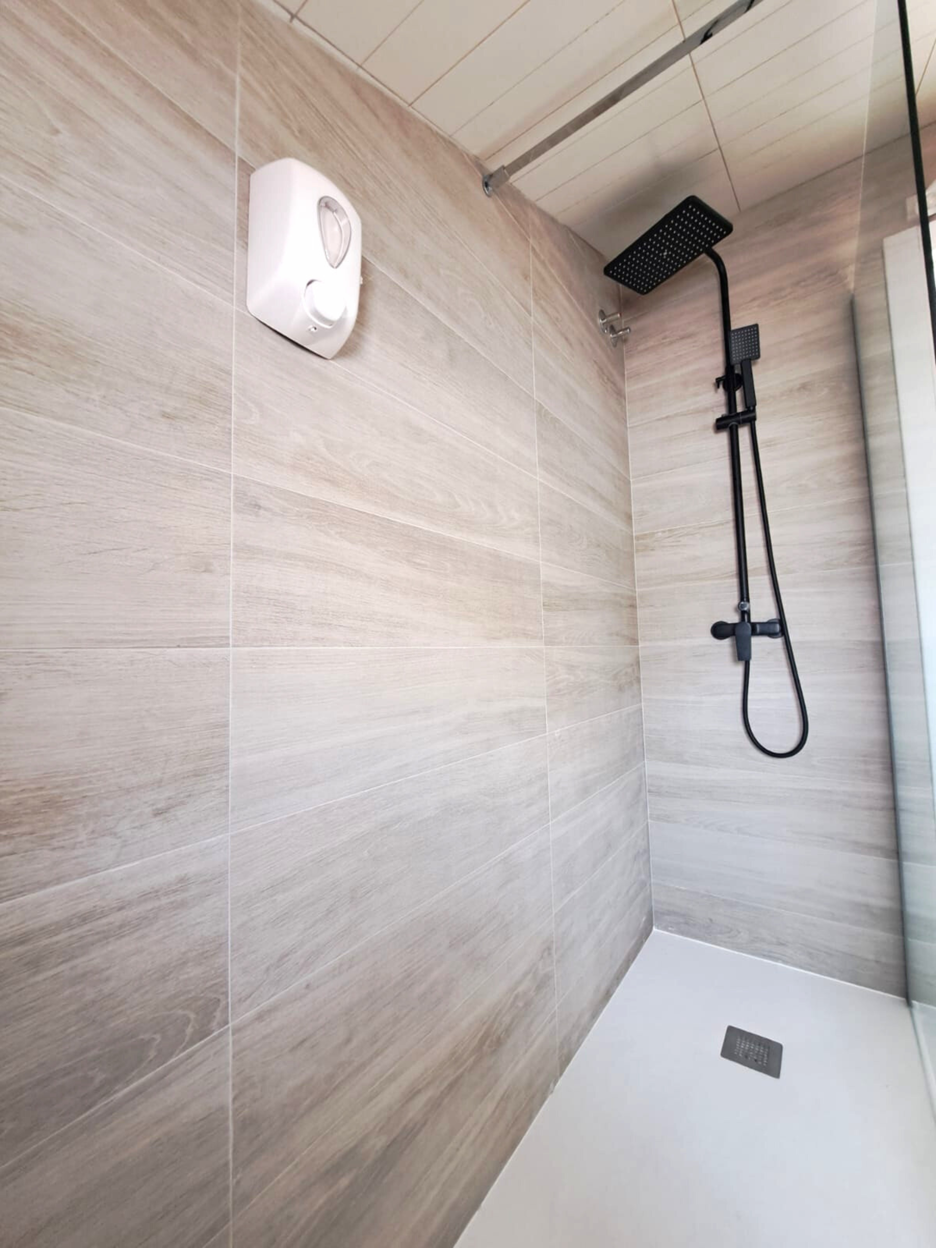 A modern shower with beige wood-look wall tiles, a black rainfall shower head, and a white soap dispenser on the wall.