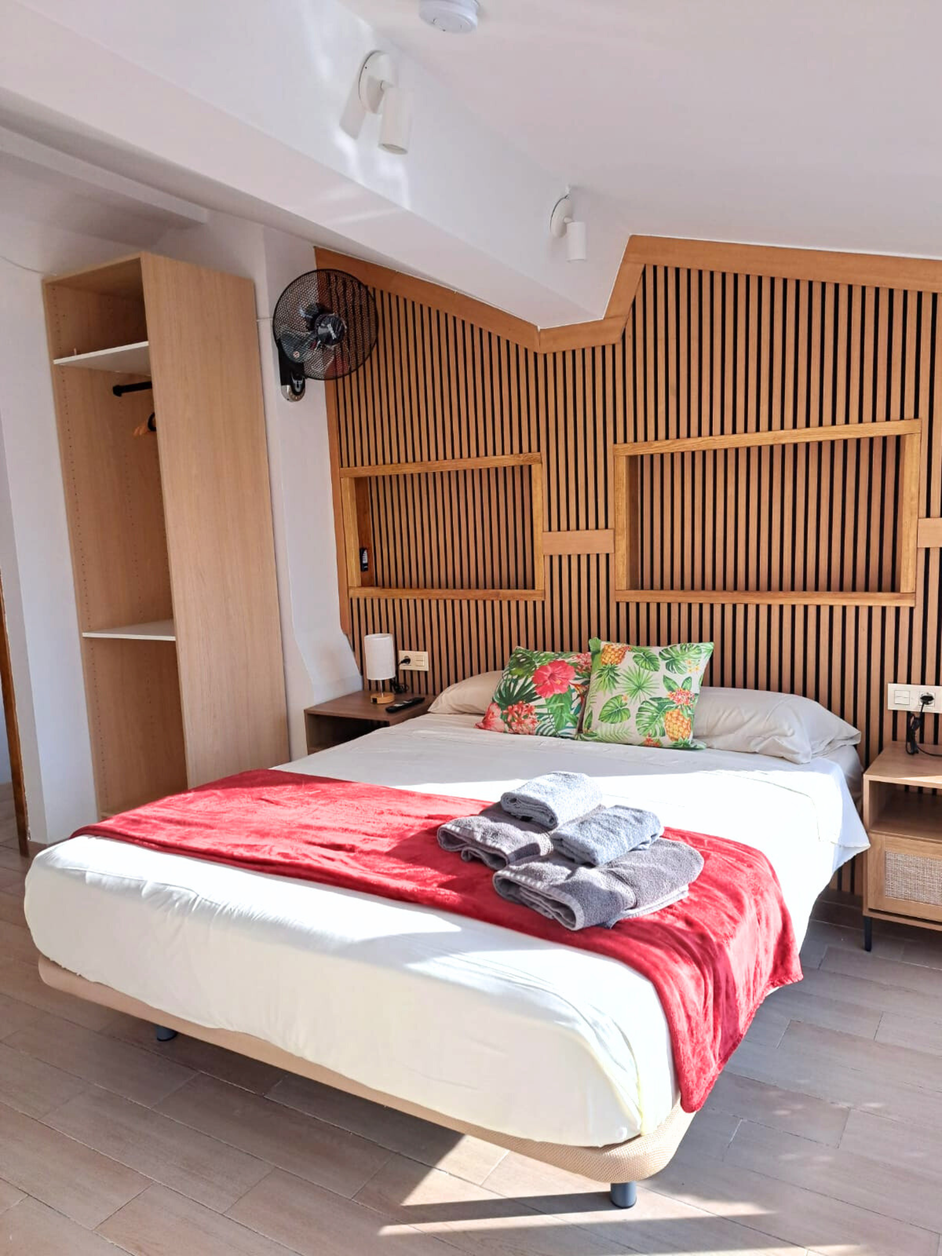 A bedroom with a bed featuring a red throw, green patterned pillows, and a wooden slat headboard, beside a tall wardrobe.