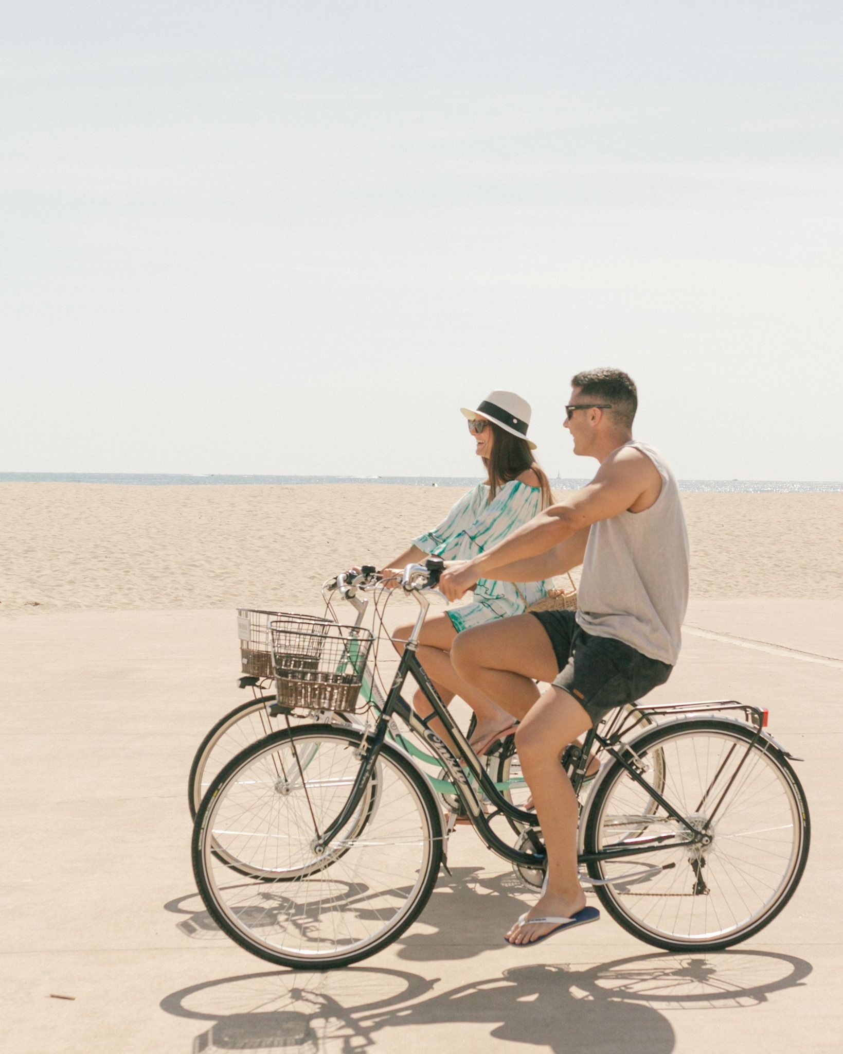 Two people ride bicycles side-by-side on a paved path along a bright, sandy beach under a clear sky.