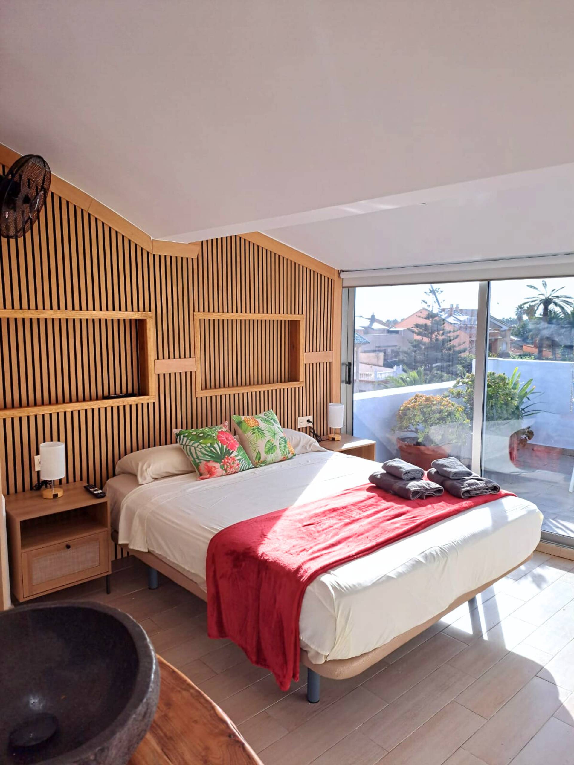 Modern bedroom featuring a bed with white linens and a red throw, a textured wooden accent wall, and a large glass balcony.