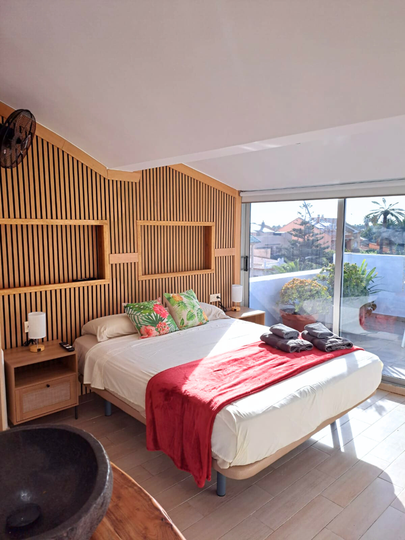 Modern bedroom featuring a bed with white linens and a red throw, a textured wooden accent wall, and a large glass balcony.